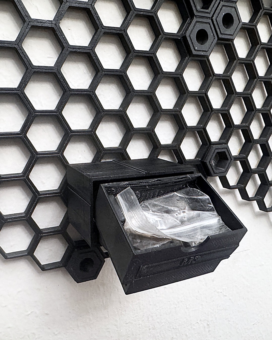 Black 3D-printed drawer pulled out of its housing on a honeycomb wall, with a bag of screws inside