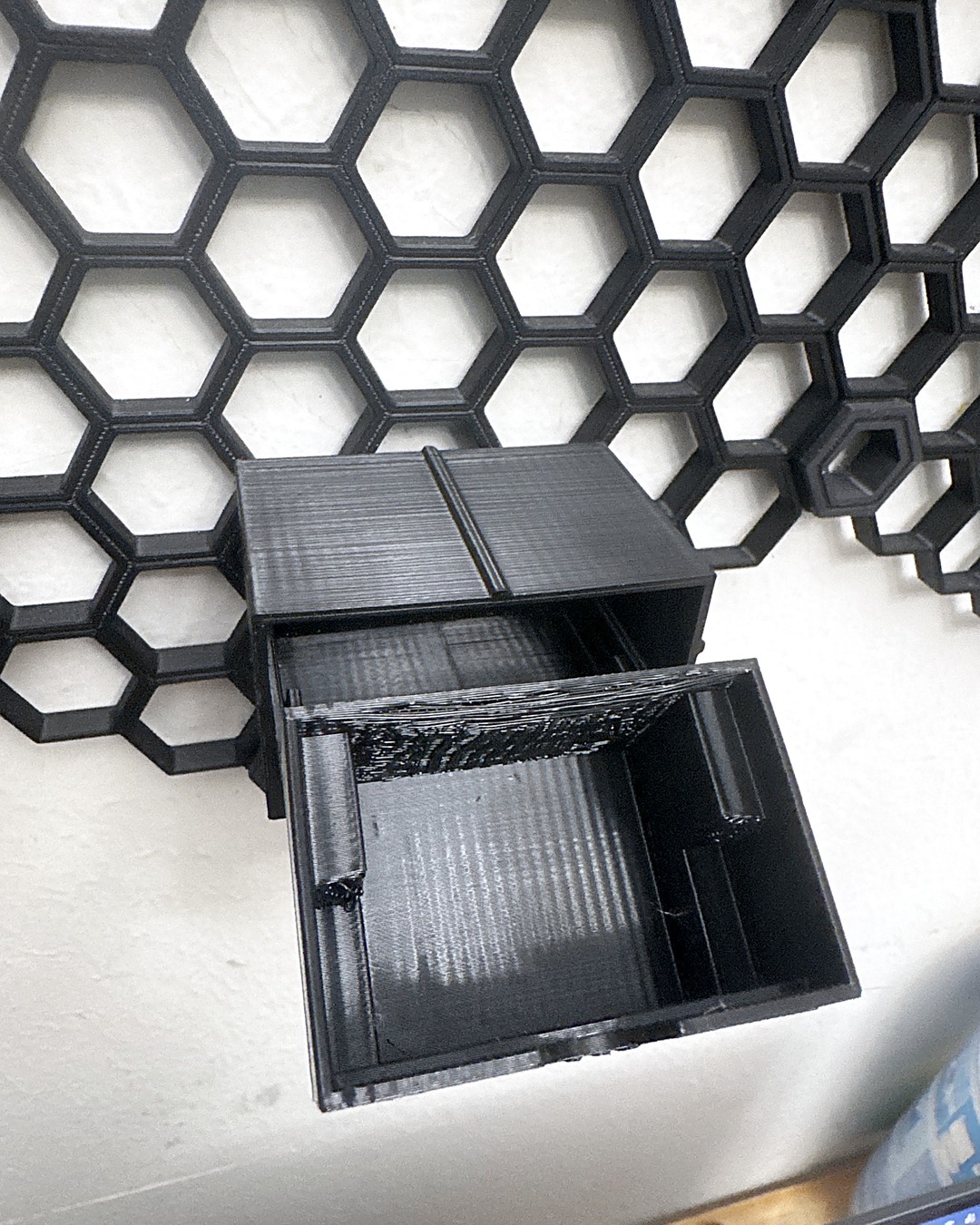 Black 3D-printed drawer pulled out of its housing and tipped forward, seen from above, on a honeycomb wall